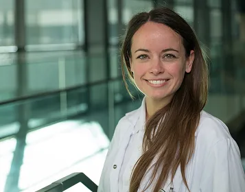 Junge Frau in einem weißen Kittel lächelt in die Kamera. Sie steht in einem modernen Gebäude mit Glaswänden und natürlichem Licht.