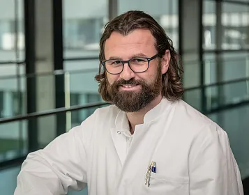 Ein lächelnder Mann mit Brille und Bart trägt einen weißen Arztkittel. Im Hintergrund sind große Fenster und ein modernes Gebäude sichtbar.