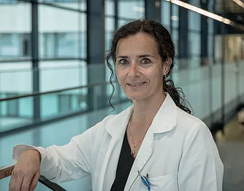 Eine Frau in einem weißen Laborkittel steht in einem modernen Gebäude mit großen Fenstern. Sie lächelt leicht und hält einen Kugelschreiber in der Brusttasche.