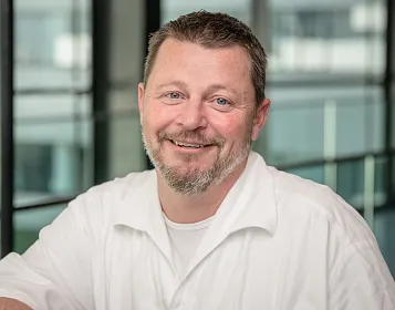 Ein Mann mit braunen Haaren und Bart lächelt in die Kamera. Er trägt ein weißes Hemd, und im Hintergrund sind Fenster mit unscharfem Ausblick sichtbar.