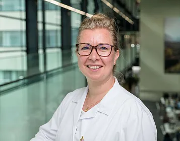 Eine Frau mit Brille und blondem Haar lächelt freundlich. Sie trägt einen weißen Kittel und steht in einem modernen Gebäude mit Glaswänden.