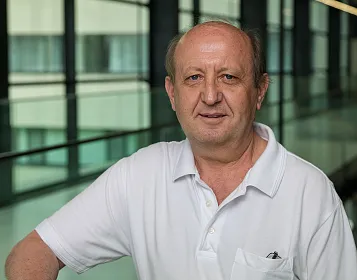 Ein Mann mittleren Alters in einem weißen Polohemd steht vor einer modernen Glasfassade. Er hat kurzes Haar und blickt freundlich in die Kamera.