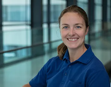 Eine Frau in einem blauen Polohemd lächelt in die Kamera. Sie befindet sich in einem modernen Gebäude mit großen Fenstern im Hintergrund.