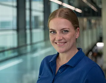 Eine Frau mit braunen Haaren trägt ein blaues Polohemd und lächelt in die Kamera. Im Hintergrund ist ein modernes Bürogebäude mit Glaswänden zu sehen.