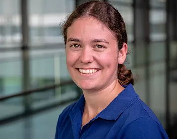 Eine lächelnde Person mit braunen Haaren in einem blauen Hemd steht in einem modernen Gebäude mit Glaswänden im Hintergrund.