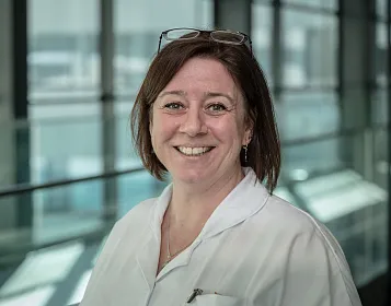 Eine lächelnde Frau in Weißkittel mit Brille auf dem Kopf steht in einem modernen Gebäude mit Glaswänden und Blick auf den Innenhof.