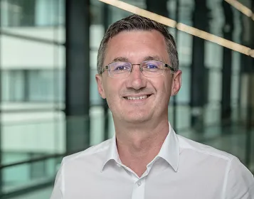 Ein Mann in einem weißen Hemd lächelt in die Kamera. Er trägt eine Brille und steht vor einem modernen, verglasten Hintergrund mit viel natürlichem Licht.