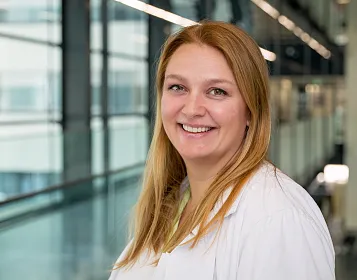 Eine lächelnde Person mit langen, blonden Haaren trägt einen weißen Laborkittel. Der Hintergrund zeigt ein modernes Gebäude mit Glaswänden und Fluren.