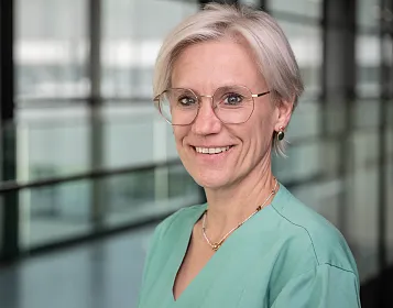 Eine medizinische Fachkraft mit Brille und kurzem, blondem Haar trägt einen grünen Kittel und lächelt. Im Hintergrund sind Glaswände zu sehen.