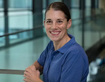 Eine Frau mit dunklem Haar und einem blauen Poloshirt steht innen in einem modernen Gebäude, lehnt an einem Geländer und lächelt in die Kamera.