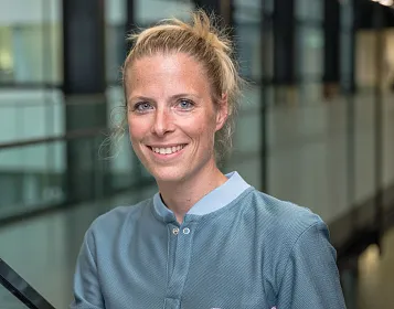 Porträt einer lächelnden Person mit blonden Haaren, die zu einem lockeren Dutt gebunden sind. Sie trägt eine graue Bluse und steht in einem modernen Gebäude.