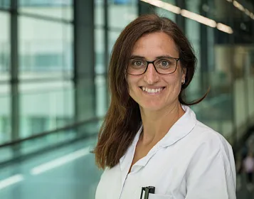 Eine Person in einem weißen Kittel und Brille lächelt in einem modernen Gebäude mit großen Fenstern und Glaswänden. Der Hintergrund ist unscharf.