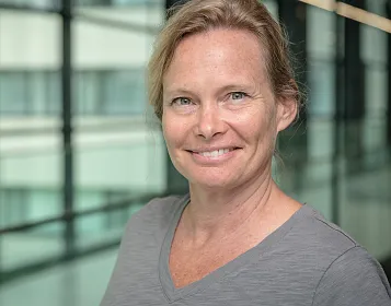 Eine lächelnde Frau mit blondem Haar und grauem Shirt steht vor einer Glaswand in einem modernen Gebäude, das viel Tageslicht hereinlässt.