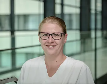 Eine Frau mit Brille und hellblondem Haar trägt ein weißes Oberteil. Sie lächelt in die Kamera. Im Hintergrund sind Glaswände und ein modernes Gebäude.