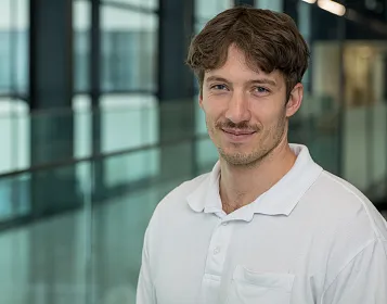 Ein Mann mit braunen Haaren und Oberlippenbart in einem weißen Polohemd steht vor einer modernen Glasarchitektur und lächelt leicht in die Kamera.