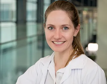 Eine lächelnde Frau in einem weißen Laborkittel steht in einem modernen Gebäude. Im Hintergrund sind große Fenster und verschwommene Konturen zu sehen.