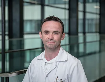 Ein Mann in einem weißen Kittel steht vor einer modernen Glasfassade in einem Gebäudeflur. Er lächelt leicht und hat einen professionellen Ausdruck.