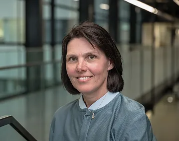 Eine Frau mit dunklen Haaren und einem blauen Umhang lächelt in einer modernen Büro-Umgebung mit Glaswänden und Beleuchtung im Hintergrund.