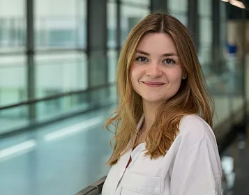 Eine lächelnde Frau mit langen, glatten Haaren steht in einem modernen Gebäude mit Glaswänden. Sie trägt ein weißes Oberteil und schaut freundlich in die Kamera.