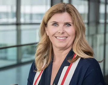 Eine lächelnde Frau mit langen blonden Haaren trägt einen dunklen Blazer. Sie steht in einem modernen, lichtdurchfluteten Gebäude mit Glaswänden.