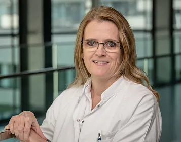 Eine Frau in einem weißen Arztkittel steht lächelnd in einem modernen Gebäude mit Glaswänden. Sie trägt eine Brille und hat schulterlanges, blondes Haar.