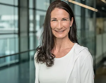 Eine lächelnde Frau mit langen, dunklen Haaren steht in einem modernen Gebäude mit Glaswänden. Sie trägt ein weißes Oberteil und eine weiße Jacke.