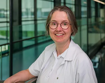 Eine lächelnde Frau mit Brille, die einen weißen Kittel trägt, steht in einem modernen, lichtdurchfluteten Gebäude mit Glaswänden.