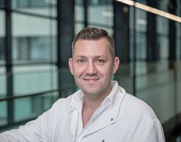 Ein Mann in weißem Kittel steht lächelnd in einem modernen Bürogebäude mit Glaswänden. Sein kurzes, dunkles Haar ist ordentlich frisiert.