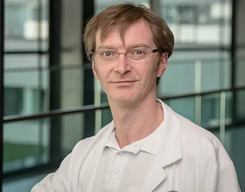 Ein Mann mittleren Alters mit Brille und braunem Haar trägt einen weißen Laborkittel. Der Hintergrund zeigt ein modernes Gebäude mit großen Fenstern.