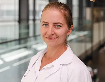 Eine lächelnde Person in einem weißen Laborkittel steht vor einer modernen Glasfassade. Das natürliche Licht betont die ruhige und freundliche Atmosphäre.