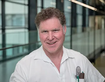 Ein lächelnder Mann in weißem Hemd steht in einem modernen Innenraum mit Glaswänden. Er trägt Stifte in der Brusttasche und hat lockiges, dunkles Haar.