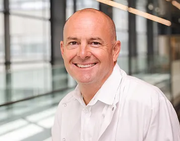 Ein lächelnder Mann in einem weißen Kittel steht in einem modernen Gebäude mit Glaswänden. Er hat kurzgeschnittene Haare und der Hintergrund ist verschwommen.