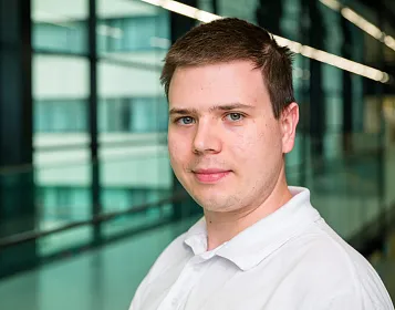 Ein junger Mann in einem weißen Hemd steht in einem modernen Gebäude mit Glaswänden. Der Hintergrund zeigt unscharf mehrere Flure und Büros.