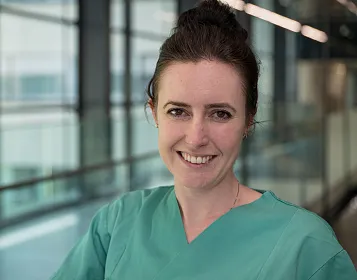 Eine lächelnde Person in grüner medizinischer Kleidung steht in einem modernen, gut beleuchteten Flur mit Glaswänden. Ihr Haar ist hochgesteckt.