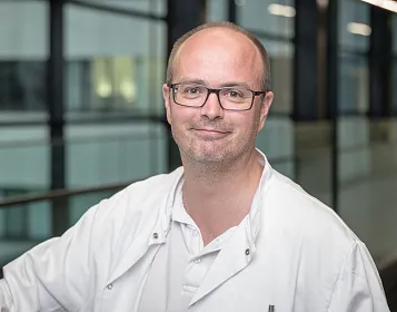 Ein Mann mit Brille und weißem Kittel steht vor einer gläsernen Wand im Inneren eines modernen Gebäudes. Er lächelt freundlich in die Kamera.