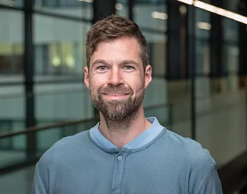 Ein Mann mit Bart und kurzem Haar lächelt in die Kamera. Er trägt ein blaues Henley-Shirt. Im Hintergrund sind unscharfe Bürogebäude zu sehen.
