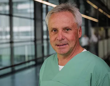 Ein Mann mittleren Alters in grüner Krankenhauskleidung steht in einem modernen Gebäude mit Glaswänden und schaut in die Kamera.