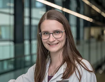 Eine Frau mit langen Haaren und Brille lächelt. Sie trägt einen weißen Laborkittel. Im Hintergrund sind Glaswände und moderne Gebäudearchitektur zu sehen.