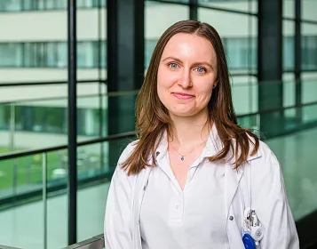 Eine Frau in einem weißen Kittel steht in einem modernen Gebäude mit Glaswänden. Sie lächelt leicht und hat langes braunes Haar.