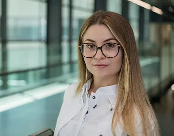 Junge Frau mit Brille und langen Haaren, trägt eine weiße Bluse. Sie steht in einem modernen Gebäude mit Glaswänden und schaut in die Kamera.