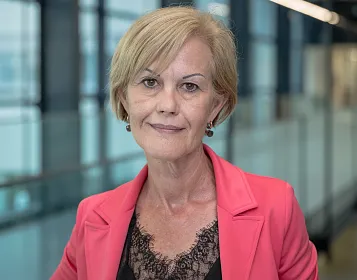 Eine Frau mittleren Alters mit kurzem, blondem Haar trägt einen pinken Blazer über einem schwarzen Spitzenoberteil. Sie steht in einem modernen Gebäude mit Glaswänden.