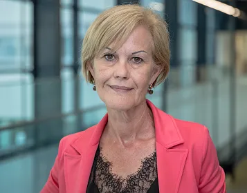 Eine Frau mit kurzem blondem Haar und einem pinkfarbenen Blazer steht vor einer Glaswand. Ihr Ausdruck ist freundlich und professionell.