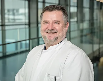 Ein lächelnder Mann mittleren Alters in einem weißen Arztkittel steht vor einer modernen Glasfassade. Er hat graues Haar und einen gepflegten Bart.