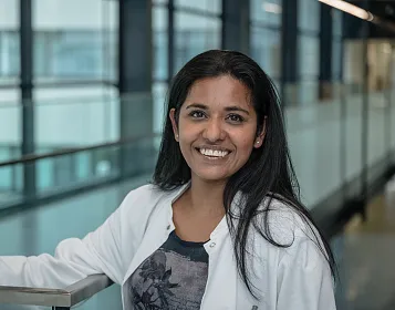 Woman in a white coat, smiling in a modern hallway with glass panels and soft lighting, exuding professionalism and warmth.