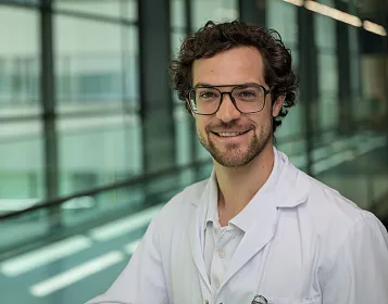 Ein lächelnder Mann mit lockigem Haar und Brille trägt einen weißen Laborkittel. Im Hintergrund sind Glaswände und ein modernes Gebäude zu sehen.