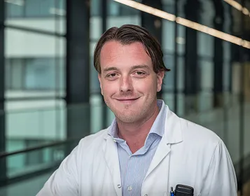 Ein Mann in einem weißen Arztkittel lächelt in die Kamera. Er steht in einem modernen Gebäude mit großen Fenstern und Glaswänden.