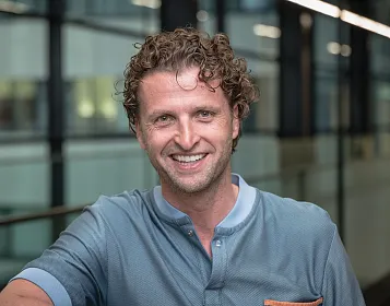 Ein Mann mit lockigem Haar lächelt in die Kamera. Er trägt ein hellblaues Hemd mit Knopfleiste und steht vor einem Hintergrund aus Glas und Metallstrukturen.