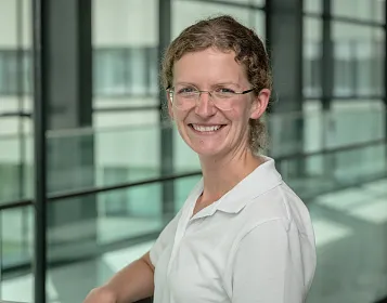 Eine Person mit Brille lächelt in die Kamera. Sie trägt ein weißes Hemd. Im Hintergrund sind große Fenster und ein modernes Gebäude zu sehen.