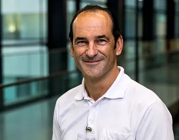 Ein lächelnder Mann mittleren Alters in einem weißen Hemd steht in einem modernen Gebäude mit Glaswänden und einer hellen, offenen Atmosphäre.