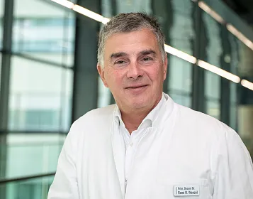 Ein lächelnder Mann mittleren Alters in einem weißen Arztkittel steht in einem modernen Gebäude mit großen Fenstern.